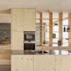 Cocina de madera maciza de abeto en color natural claro, compuesta por un módulo con puertas donde está integrado el horno y el microondas. Al lado derecho hay una estantería formada por estantes horizontales y verticales de madera de abeto. También aparece una isla de cocina con puertas de armario y donde se encuentra el fregadero y la vitrocerámica.